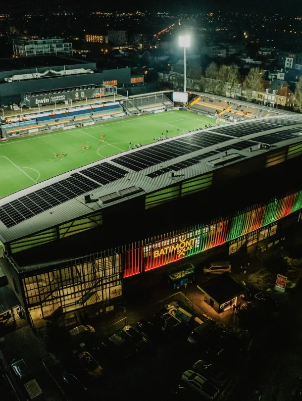 Luchtbeeld bij avond van een voetbalstadion waarvan de tribunedaken volledig met zonnepanelen zijn belegd; de gevel draagt het logo van Batimont Solar