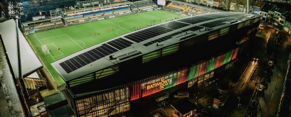 Luchtbeeld bij avond van een voetbalstadion waarvan de tribunedaken volledig met zonnepanelen zijn belegd; de gevel draagt het logo van Batimont Solar